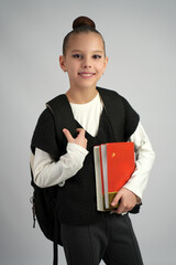 Happy young girl in school uniform holding books and wearing a black backpack, posing against a...