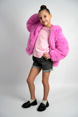 Close-up of a smiling girl in a fluffy pink jacket and sunglasses on her head, framing her face with her hands against a grey backdrop