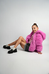 A stylish girl wearing a bright pink faux fur jacket and pink shirt crouching and smiling against a clean grey studio background