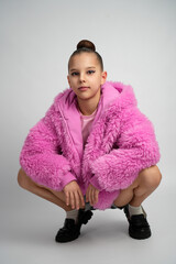 A stylish girl wearing a bright pink faux fur jacket and pink shirt crouching and smiling against a clean grey studio background