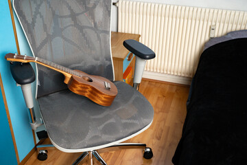 Brown wooden ukulele resting on a gaming chair next to a bed in a casual home interior. Everyday leisure in a bedroom setup. Concept of home, lifestyle, music, leisure, casual.