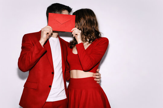 Red dressed couple hiding faces behind love letter envelope, Saint Valentine&rsquo;s Day celebration, romantic relationship concept, passion symbolism, minimal background with copy space