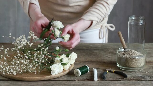 Florist at work: woman shows how to make romantic candle holder using a glass jar, carnations flowers, gypsophila paniculata twigs and sand. Step by step, tutorial. Images available.