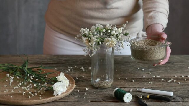 Florist at work: woman shows how to make romantic candle holder using a glass jar, carnations flowers, gypsophila paniculata twigs and sand. Step by step, tutorial. Images available.