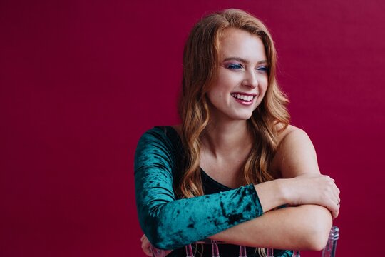 Portrait of a redheaded smiling woman wearing make-up and an off shoulder dress sitting on a chair in front of a red background 
