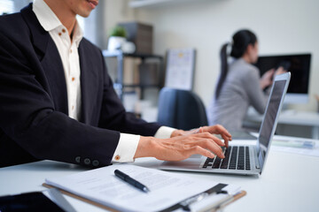 Fototapeta premium Asian business man sitting at desk, using laptop computer in open space office, female colleague is sitting and working in the back