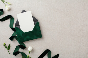 Dark green wedding envelope with invitation card, green ribbon, white flowers on stone table. Flat lay, top view.