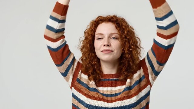 A cheerful young person with vibrant red, curly hair and a radiant smile stretches gracefully, reaching upwards with both arms against a clean, light background. Dressed in a cozy, colorful striped kn