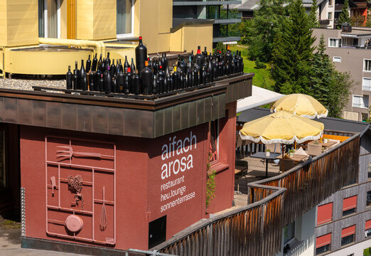 Arosa, Switzerland - August 13, 2025: A rooftop Aifach Arosa restaurant terrace with black bottles arranged decoratively, featuring a restaurant sign, outdoor seating with umbrellas, and green trees i