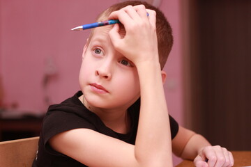 Obraz premium The middle plan of a teenager sitting at a table at home: he leans his elbow on the table and holds a pen to his temple, as if he is considering a task or pauses in his work. It is suitable for illust