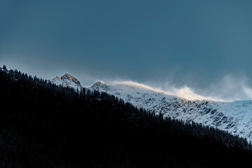 Wind blows snow across a mountain ridge, backlit by the sun, creating a glowing effect as the snow...