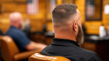 A man with a neatly trimmed hairstyle sits in a barbershop, with a rustic wooden interior and another customer in the background.