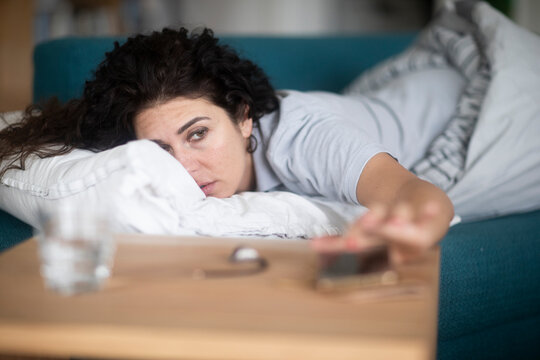 Sleepy Woman lying on a sofa with a duvet turning off the alarm on her mobile phone