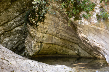 Fototapeta premium Natural rock formation with shallow water pool inside stone cavity. Layered limestone walls and moss vegetation in shaded outdoor environment