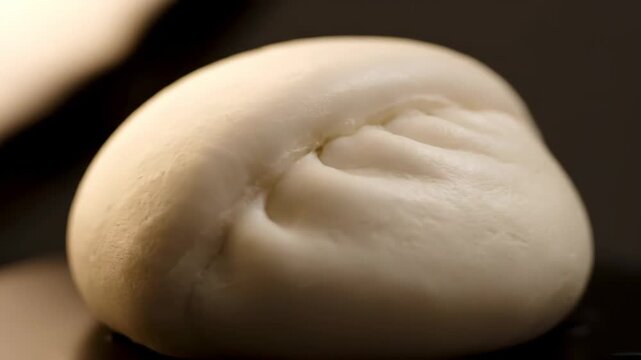 Extreme close-up of a perfectly steamed white Asian bun (Baozi or Mantou) showcasing the delicate pleating and soft texture against a dark, minimalist background.