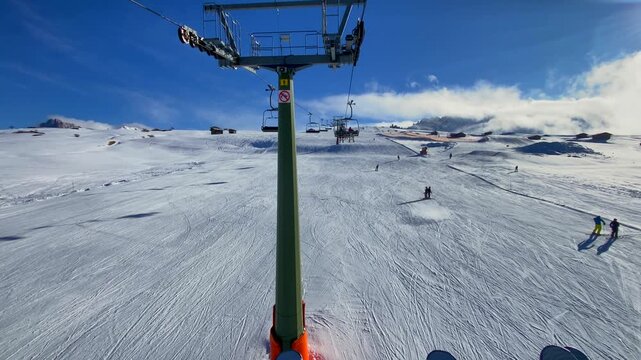 A wide-angle shot of skiers on a chairlift at Alpe di Siusi in the Dolomites. A panoramic shot of the lift ascending the snow-covered slopes of the Seiser Alm. Winter sports in the Italian Alps skiers