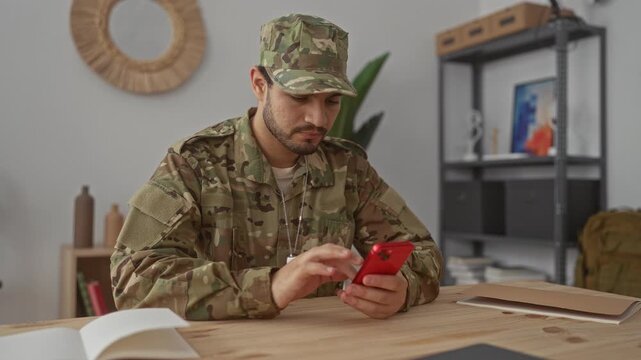 Young hispanic man in camouflage uniform taps red smartphone at wooden desk in building; connection.