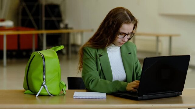 Young woman in green blazer and glasses working on dell laptop in classroom with bright backpack on desk, concept serious female student studying online and doing homework.