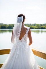 Fototapeta premium Elegant bride in white lace wedding dress looking at a calm lake from a wooden pier