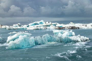 Large icebergs near iceland diamond beach glacier drift calmly in a lagoon surrounded by water and mountains. The skies are gray with clouds, and a few birds fly overhead. © stockphoto-graf