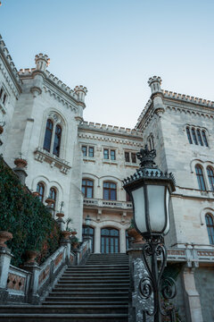 View of a grand castle with stone steps leading up to its ornate facade, framed by an antique lamp post against a clear sky, Grignano Miramare, Friuli-Venezia Giulia, Italy.