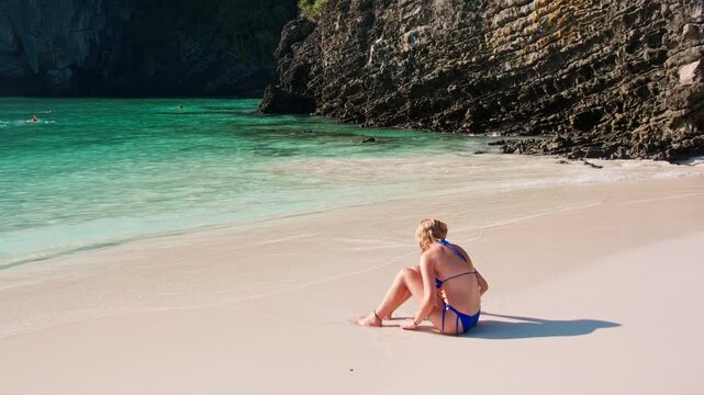 Woman in a swimsuit sitting on the sand of Nui Beach in Krabi, Thailand, enjoying the serene ocean view with limestone karsts in the background and longtail boats floating on the turquoise water