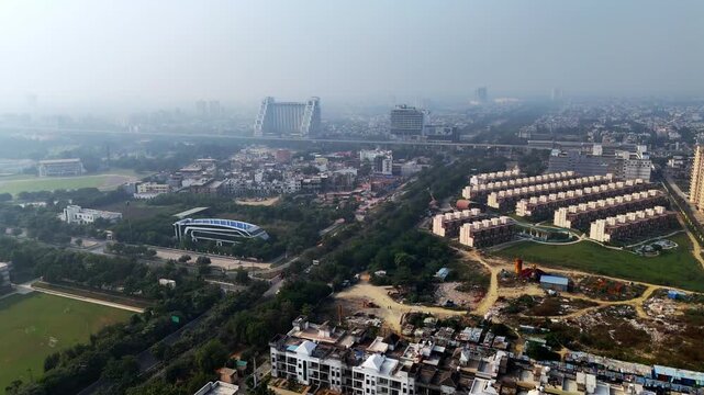 Aerial view of Greater Noida under light haze, showing residential housing clusters, green corridors, ongoing construction, and elevated road connectivity highlighting rapid urban expansion.