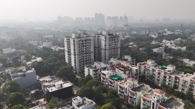 Aerial video of Sector 36, RHO I, Greater Noida mid-rise and high-rise residential apartments, surrounded by dense tree cover, with distant buildings softened by winter haze and air pollution.