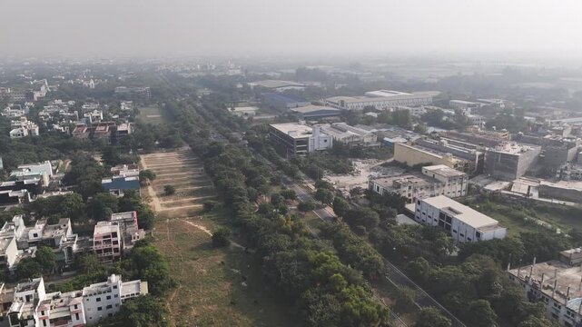Aerial view of an industrial and institutional corridor, with tree-lined roads, factory buildings, warehouses, and light traffic stretching into the hazy urban horizon.