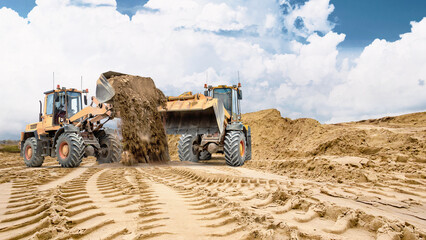 Two wheel loaders. Heavy equipment is working on sandy soil at a construction site. Track marks are visible in the sand, and a large pile of sand is visible in the background © Anoo