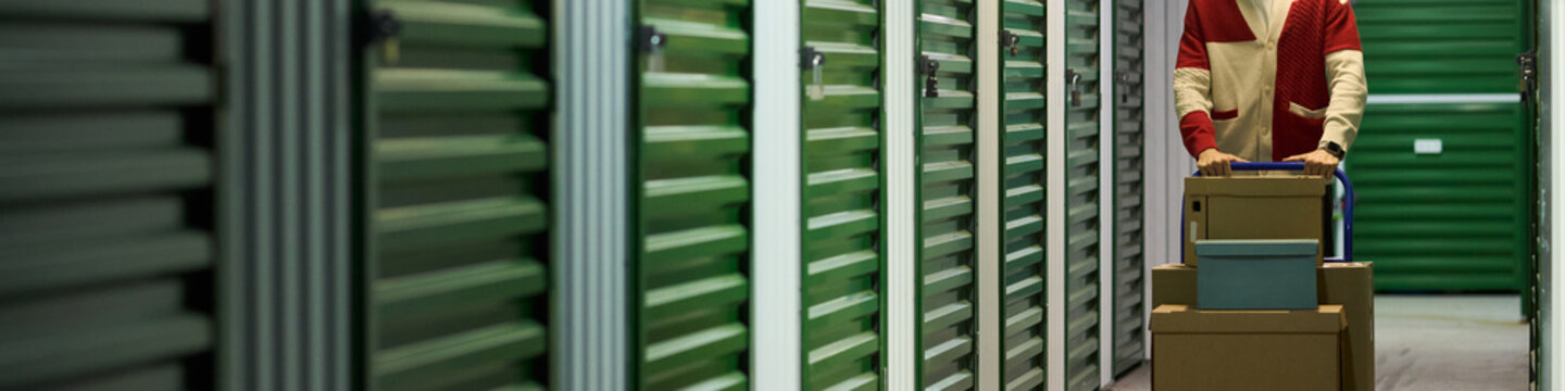 Middle aged Caucasian man pushing stack of cardboard boxes on dolly cart through hallway lined with green storage unit doors, focusing on transporting belongings in self storage facility