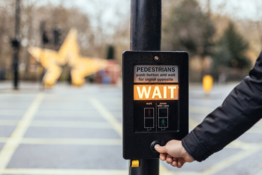 Pedestrian pressing WAIT button at crosswalk on street with holiday lights in the background.