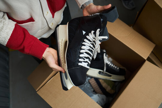Caucasian young adult man holding pair of black ice hockey skates above open cardboard box, preparing for storage or packing, hands and forearms visible, indoor setting