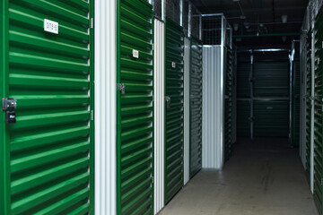 Row of green metal storage unit doors with padlocks lining corridor in self storage facility, empty hallway extending into darkness, industrial environment with secure access