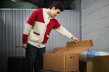 Young adult Caucasian man standing in storage facility opening cardboard box, examining contents with focused expression, black suitcase and packed items visible in background