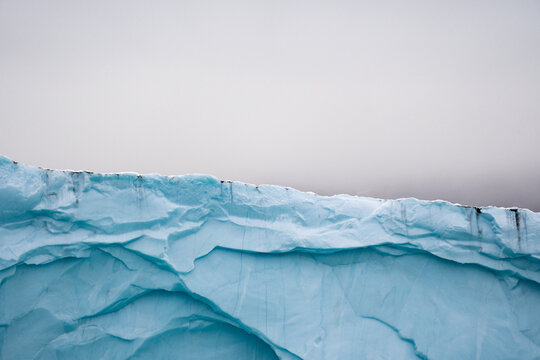 View of a massive, pale blue iceberg, its jagged edges cutting a stark line against the muted grey sky above, Scoresby Sund, Greenland.