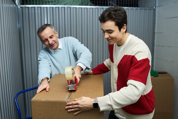 Caucasian middle aged man and Caucasian teenager boy sealing cardboard box with packing tape dispenser in storage facility, both smiling and working together on moving or organizing belongings