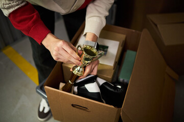 Young adult Caucasian man packing cardboard box with personal belongings, holding small trophy in...