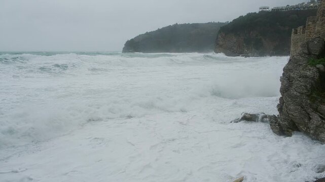 Rough stormy sea with powerful waves crashing against rocky coastline and cliff under overcast sky. Viewpoint at Rock Beach, Budva, Montenegro