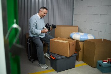 Middle aged Caucasian man sitting on suitcase holding boxing gloves in storage unit, surrounded by cardboard boxes and plastic bin appearing to organize or pack belongings