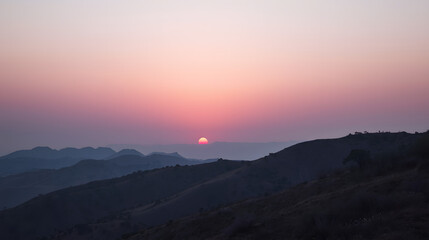 Fototapeta premium Sunset over the Mountains: The sun dips below the horizon, painting the sky with fiery hues, silhouetting the rugged mountain range in a display of natural grandeur.