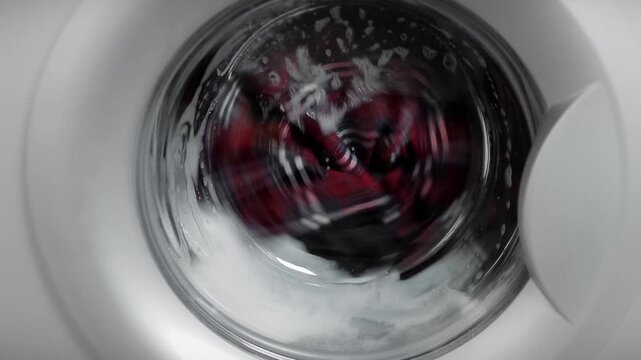 Washing machine glass door window with spinning and rotating clothes inside with water splashes and foam of laundry detergent. Full HD video with audio