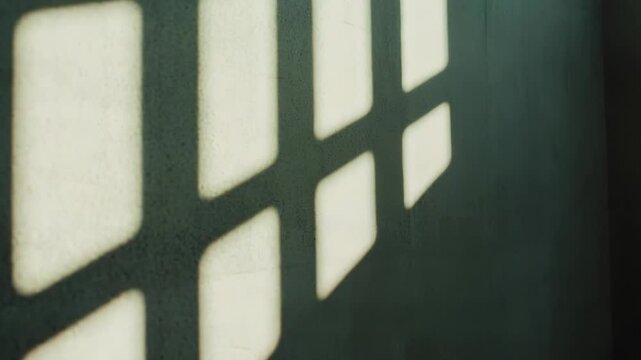 Bright sunlight streams through a window grid, casting bold geometric squares and sharp shadows across a dark textured wall, minimalist contrast of light and form