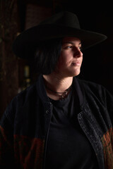 Cowgirl in a wide hat gazes into the distance at a dimly lit ranch setting