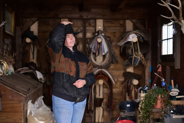 Fototapeta premium Cowgirl in a rustic barn adjusts her hat among saddles and tack, ready for work