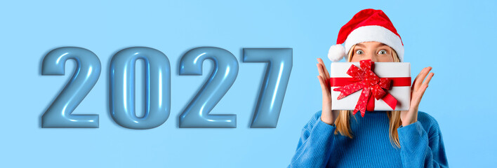 Obraz premium A person wearing a red Santa hat holds a gift box with a ribbon in front of their face. Large numbers display 2027 in blue, creating a festive scene for New Year celebrations.