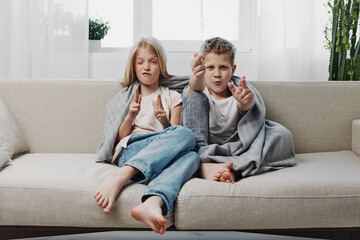 Obraz premium Two playful children sit barefoot on a beige sofa making funny faces and hand gestures in a bright living room. Concept of childhood fun, creativity, and sibling bonding.
