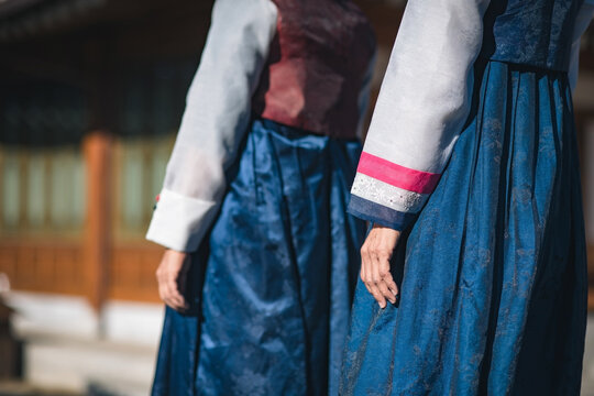 Two women wearing traditional Korean Hanbok clothing with close-up of textures and colours of sleeves and costume, walking outdoors in sunny day in a traditional house