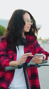 Portrait of woman dressed in red and black shirt with white t-shirt which wearing glasses and earphones on her neck looking into camera while walking with electic scooter near big urban building