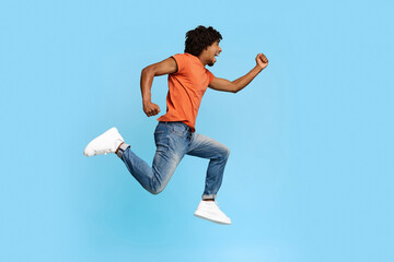 Obraz premium Emotional young african american guy in casual outfit running towards copy space on blue studio background, happy handsome millennial black man with bushy hair catching exciting deal
